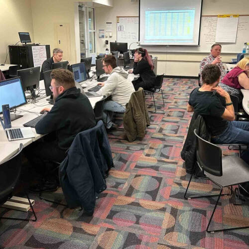 A classroom-style computer lab with nine adults seated at desks arranged along two curved rows, each working intently at desktop computers. A large projection screen at the front displays a spreadsheet, while the space is lit with overhead lights